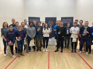 A group photo of amateur squash contestants of all genders and ages. They are posing for the photo with smiles and prizes in their hands, in front of our company banners.